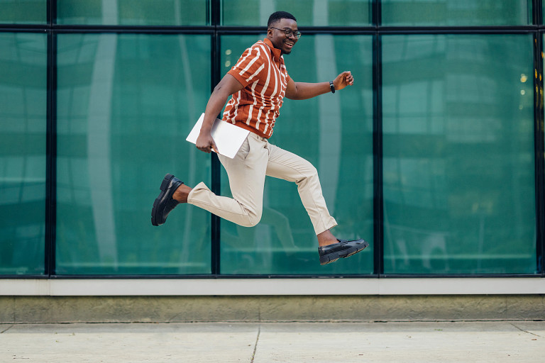 A young professional in front of a city building and holding a laptop jumping into the air